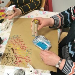wo people stamping colorful patterns on brown paper using cardboard tubes and paint trays.