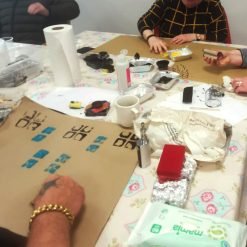 Group of people engaged in a hands-on printmaking workshop, using rollers, paint trays, and cardboard stamps on a floral-covered table with patterned brown paper sheets.