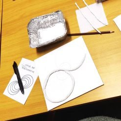 Art project in progress on a wooden table featuring a white sheet with overlapping circular outlines, one partially covered with rolled paper or clay; nearby are a pencil, aluminum tray, paper towel, and a card labeled 'SERENE AND TRANSFORMATIVE' with a pen on top