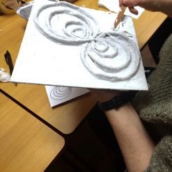 Person shaping a swirling figure-eight design with white plaster or clay on a square canvas, using a wooden stick; printed reference sheet with matching pattern partially visible on a wooden table in a classroom or workshop setting