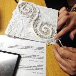 Person working on a textured white sheet with two spiral designs made of raised material and sprinkled sand; using a tool to refine the spirals on a wooden table with a printed paper about nature-inspired art partially visible underneath