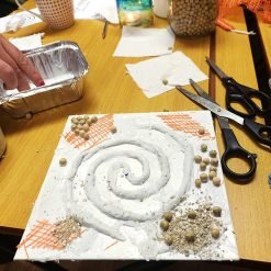 Art project on a wooden table featuring a square white base with a raised spiral design made of textured clay or plaster, decorated with beige beads, crushed sand-like material, and orange mesh fabric; surrounded by crafting tools and materials