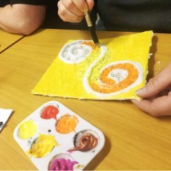 Person painting a textured canvas with a yellow background and two spiral designs—one orange and one gray—outlined in white; paint palette with warm colors and art supplies visible on the table
