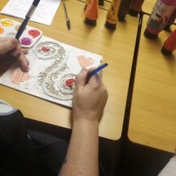 Person using a paintbrush to work on a textured canvas featuring swirling designs with granular material, red and pink painted areas, and orange crosshatched heart shapes; surrounded by paint bottles, a palette with warm colors, and a worksheet on a wooden table