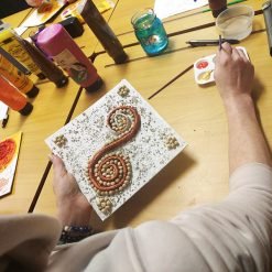 Person holding a square canvas with a textured spiral design made of beads and earthy materials, surrounded by art supplies including paint bottles, brushes, a water jar, and a palette with red and yellow paint on a wooden table