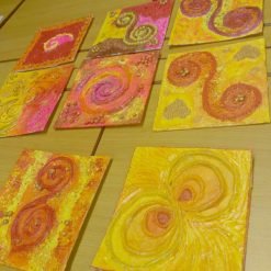 Nine square mixed-media art pieces arranged on a wooden surface, each featuring vibrant swirling spiral patterns in warm colors like yellow, orange, pink, and red, with textured elements such as beads and thick paint