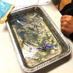 Aluminum tray with marbled swirls of blue, green, and purple ink in water, hand nearby.