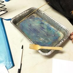 Aluminum tray with swirling gel in blue, purple, and white, wooden tools resting on top.