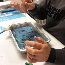 Person swirling blue, green, and purple ink in aluminum tray with wooden stick during marbling activity.