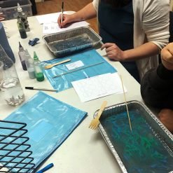 Two people marbling ink in aluminum trays with colored bottles, wooden tools, and worksheets labeled 'Test Area'.