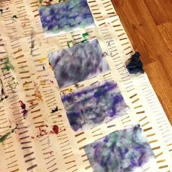 Five marbled art sheets in blue, purple, and green on paint-marked white cloth over wooden floor.