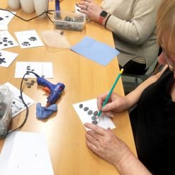 Group crafting pebble art on white cards using glue and pencils, with stone containers and tools on table.