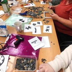 Group crafting pebble art on white cards using glue and pencils, with stone containers and tools on table.