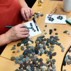 Person arranging pebbles into designs on white paper using a green pencil, with assorted stones nearby.