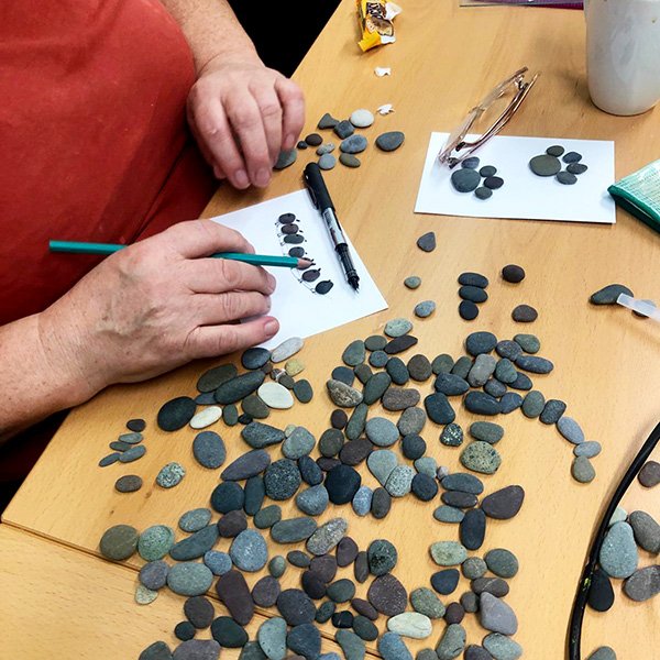 Person arranging pebbles into designs on white paper using a green pencil, with assorted stones nearby.