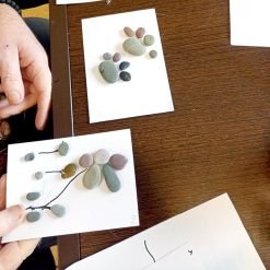Hands arranging pebbles into animal and flower designs on white cards during a tabletop craft activity.