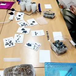 Person holding pebble art card at a table with mugs, tools, and stone containers during a craft session.