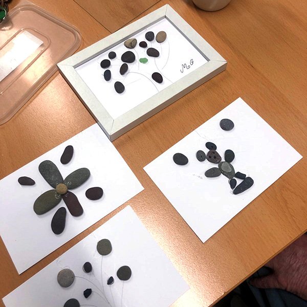 Four pebble artworks on white paper depicting trees and flowers, one framed and signed 'MG'.
