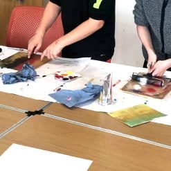 Two people using rollers and paint palettes during a printmaking activity at a covered table.