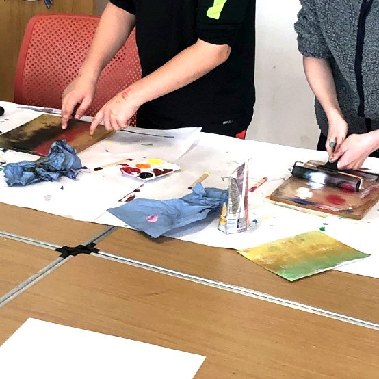 Two people using rollers and paint palettes during a printmaking activity at a covered table.