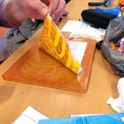 Person lifting paper from inked plate to reveal red and yellow abstract print in a creative workspace.