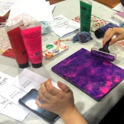 Person using roller to apply purple and pink paint on a plate surrounded by stencils and printed papers.