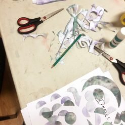 Crafting workspace with scissors, glue stick, pencil, and cut-out paper shapes representing moon phases in green and purple, arranged around a sheet labeled 'MOON PHASES