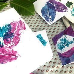 Painted leaf prints in purple, blue, and teal on white paper, surrounded by green leaves and patterns.