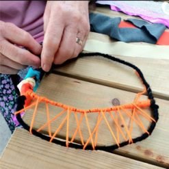 Person crafting a circular frame wrapped in black yarn with orange yarn woven in a web-like pattern.