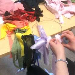A colorful fabric tassel being tied, with strips of pink, red, yellow, green, blue, and purple fabrics on a light wooden surface.