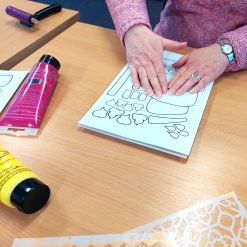 Person pressing outlined shapes on paper using stencils, paint tubes, and a roller tool.