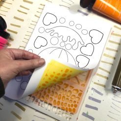 Hand lifting paper with yellow-orange patterns, revealing stencil and black outlined shapes beneath.