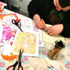 Person drawing on paper at a table with colorful patterned sheets, scissors, and markers.