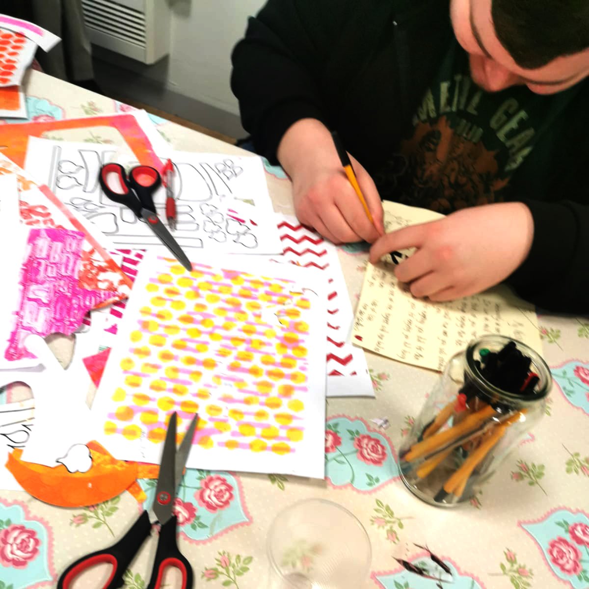 Person drawing on paper at a table with colorful patterned sheets, scissors, and markers.
