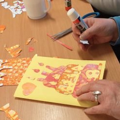 Person gluing paper cutouts onto a yellow greeting card with a birthday cake and heart design.