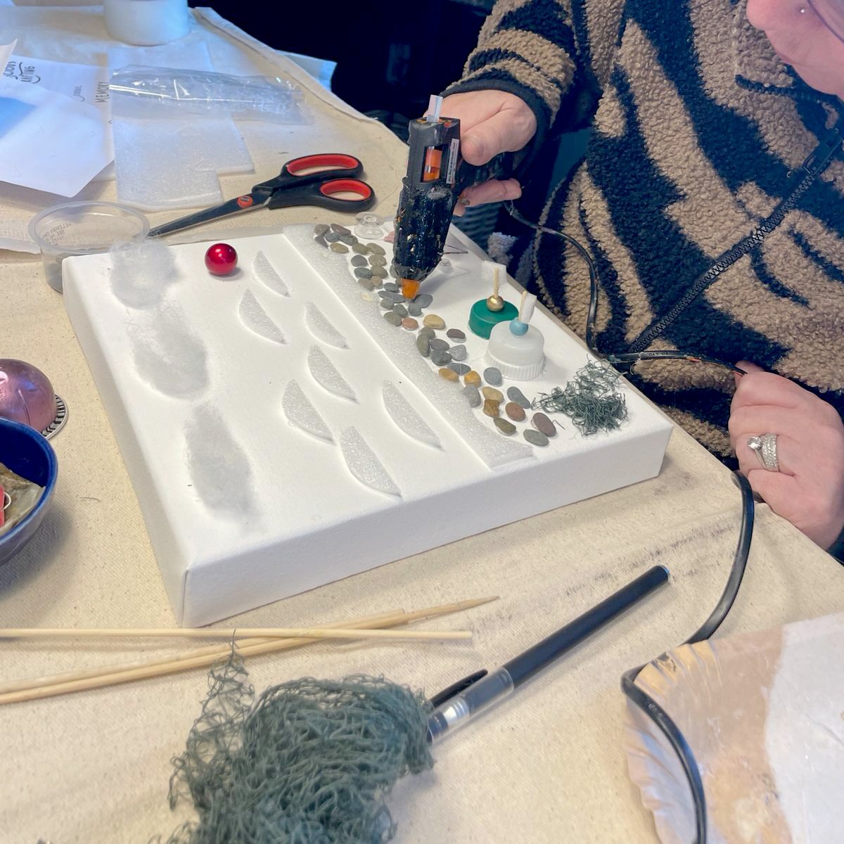 Person using hot glue gun to attach pebbles and mixed materials onto white canvas amid craft supplies.