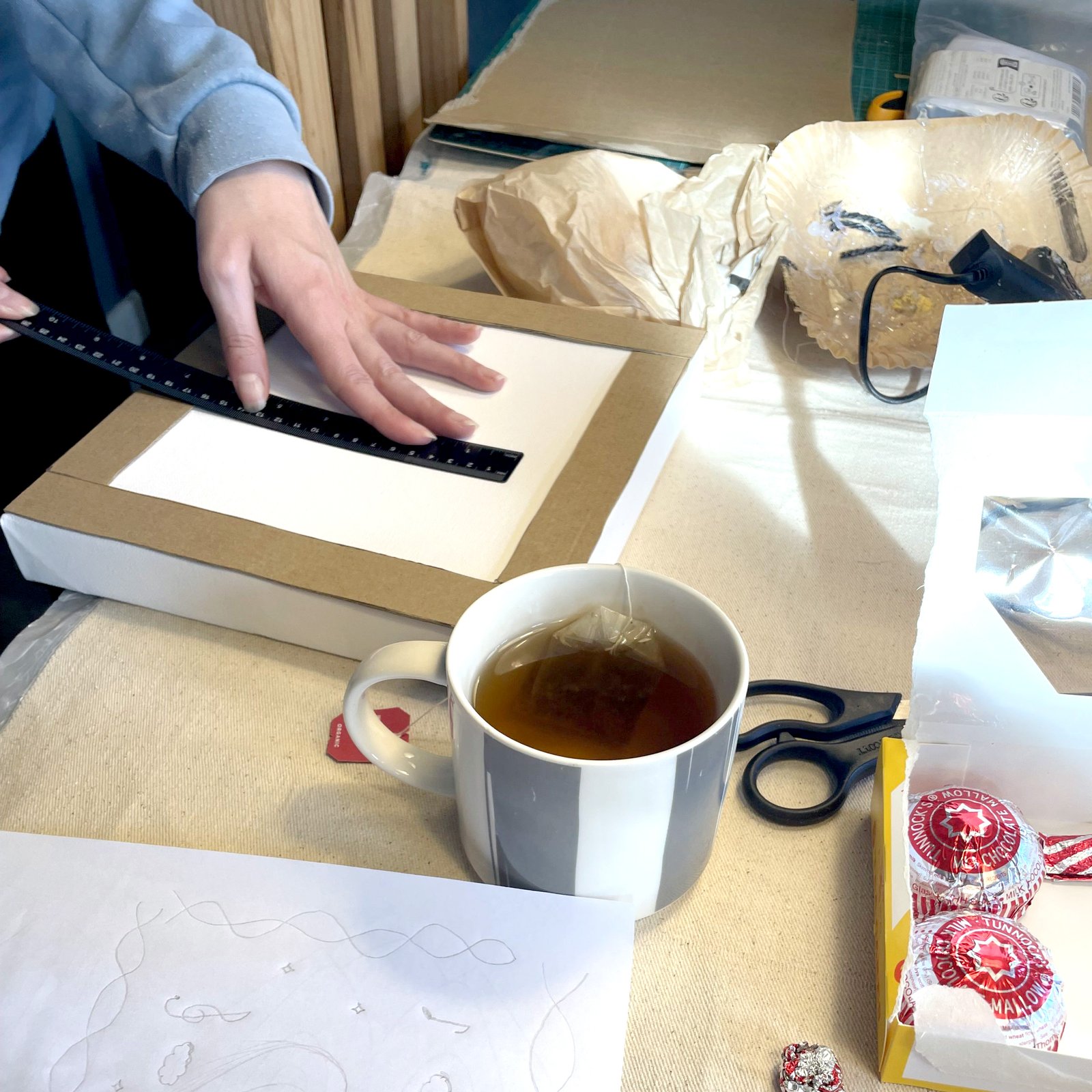 Person using ruler on white sheet amid art supplies, tea, glue gun, and Tunnock's candy box.