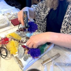 Person using hot glue gun to attach colorful mesh, yarn, and natural materials onto canvas in art project.