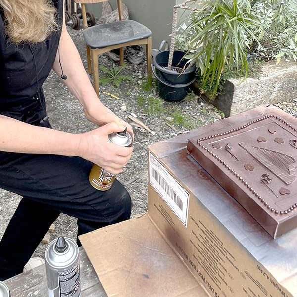 Person spray-painting embossed decorative object with Montana Gold can on cardboard box outdoors.