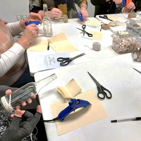 Group decorating glass bottles with twine and glue guns at craft table labeled 'Juguem a Crear'.