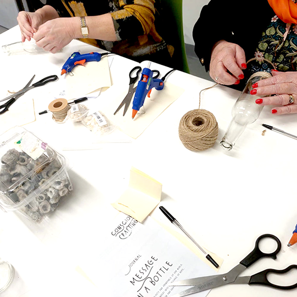 Two people crafting 'Message in a Bottle' with twine, glue guns, and glass bottles on organized table.