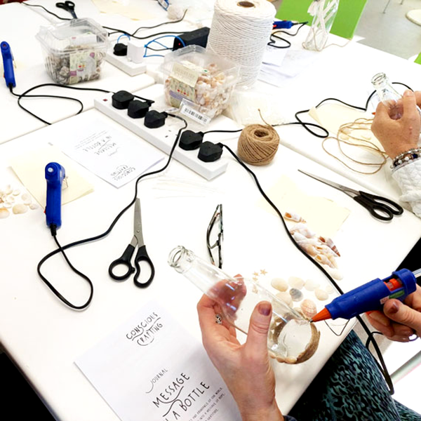 People decorating glass bottles with twine and seashells at craft table labeled 'Conscious Crafting'.