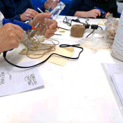 Person gluing seashells and twine onto glass bottle at craft table with glue guns and natural materials.