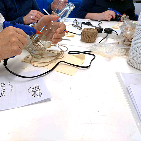 Person gluing seashells and twine onto glass bottle at craft table with glue guns and natural materials.