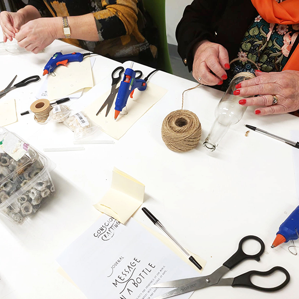 Two people crafting 'Message in a Bottle' with twine, glue guns, and small items on cluttered table.