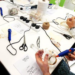 Two people decorating glass bottles with twine and seashells at craft table labeled 'Message in a Bottle'.