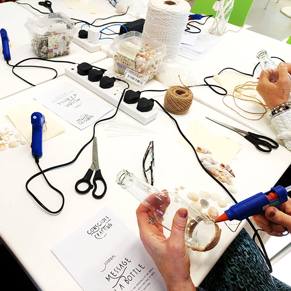 Two people decorating glass bottles with twine and seashells at craft table labeled 'Message in a Bottle'.