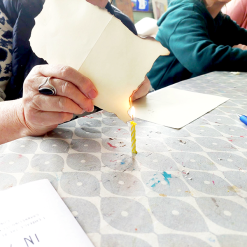 Person burning edge of paper over lit birthday candle on patterned table with art supplies.