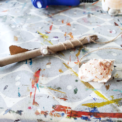 Handmade wand wrapped in twine with pearl and metal leaf beside seashell on paint-splattered surface.