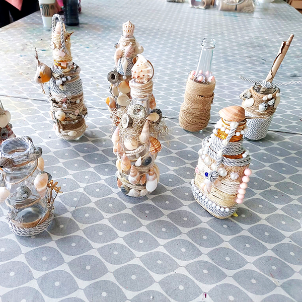 Decorated glass bottles with twine, seashells, beads, and corks arranged on patterned tablecloth.